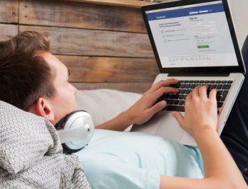 person laying in bed with headphones on, working at their laptop