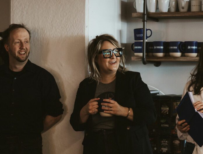 John, Lindsey and Jerri standing in front of the coffee maker and cups laughing at Gemini's staff meeting.